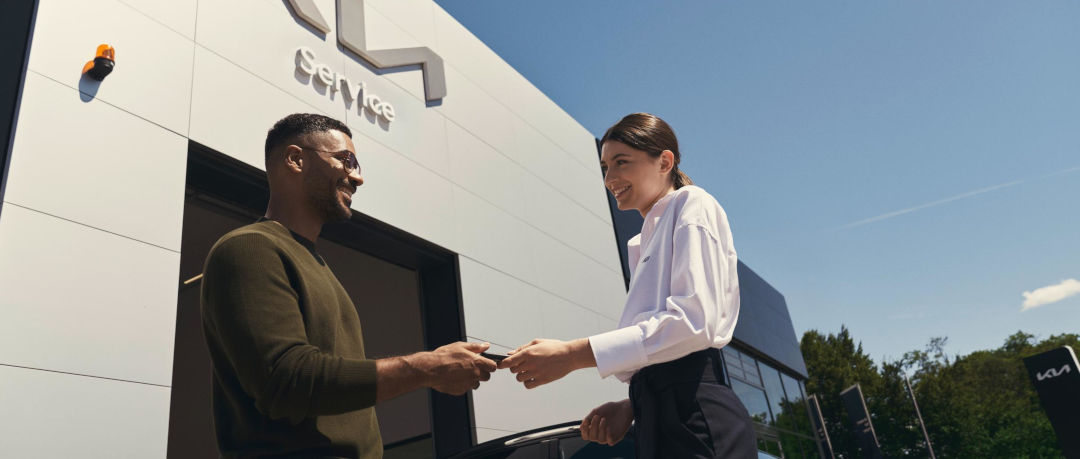 Serviceversprechen, Service-Mitarbeiter und Kundin stehen vor einer Kia Service-Werkstatt, Schlüsselübergabe