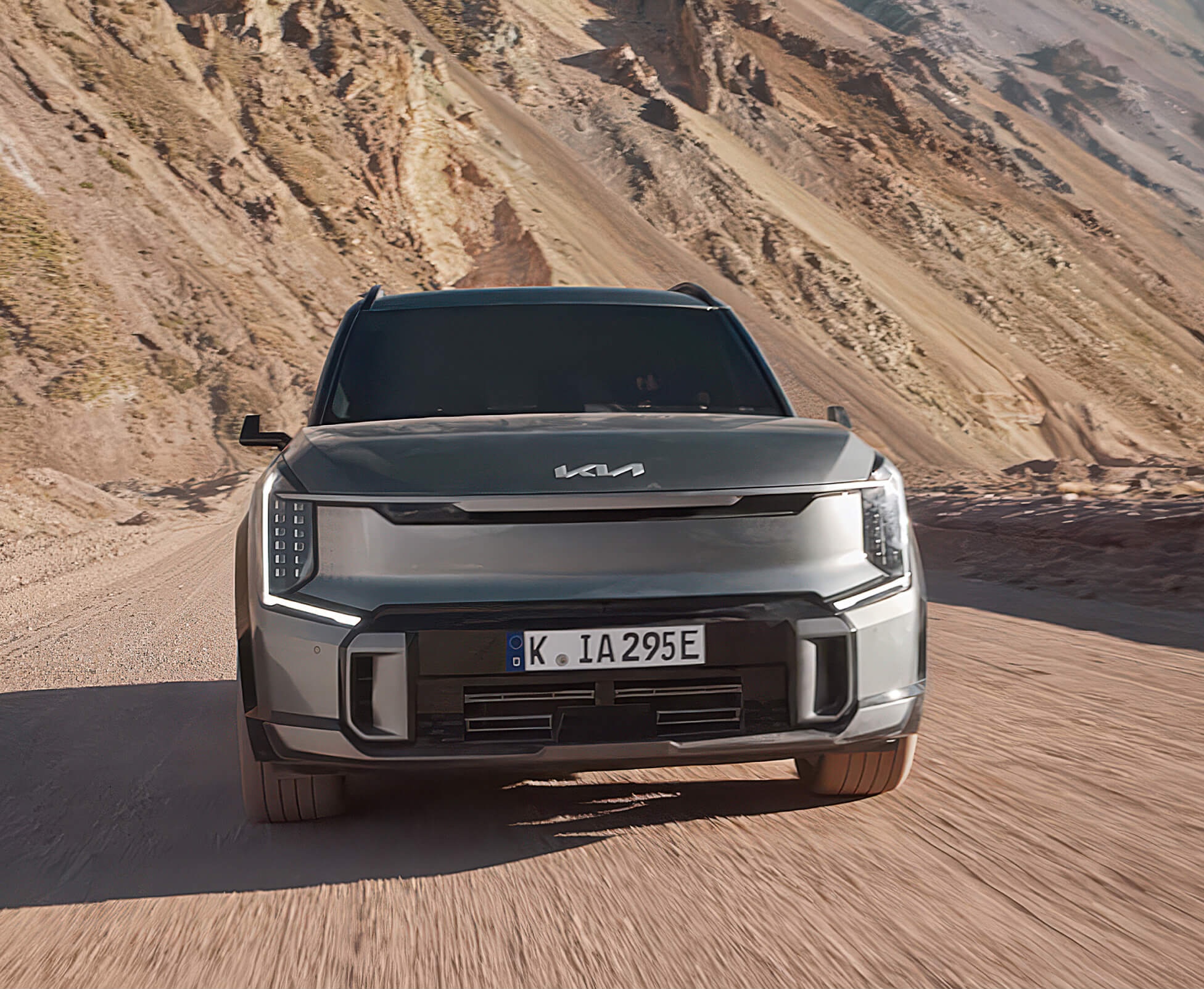Kia Soundscapes. Frontansicht eines Kia EV9 in Pebble Grey Metallic der auf einer staubigen Straße durch eine Wüstenlandschaft bergauf fährt.