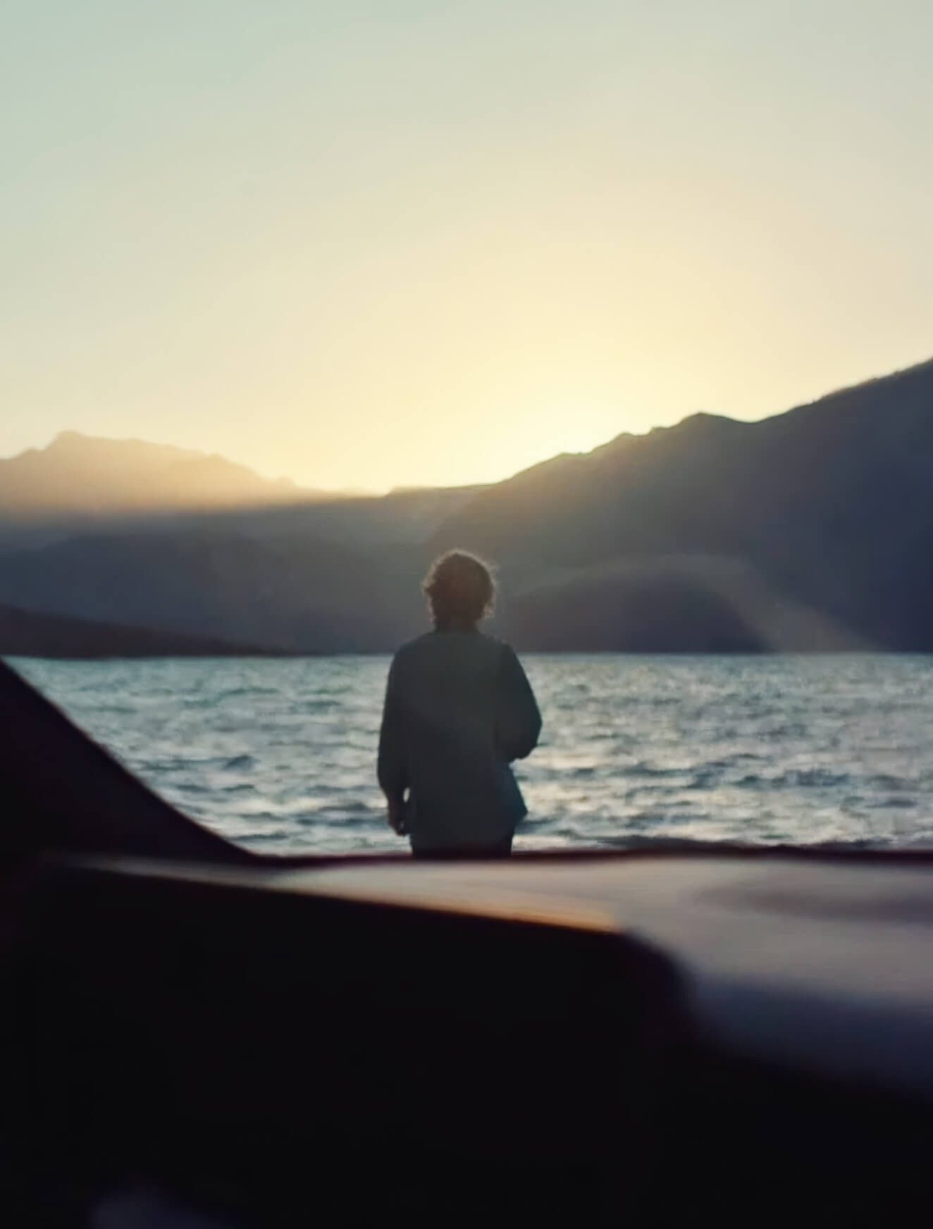 Kia Soundscapes. Blick durch das Fenster eines Kia EV9 auf einen Mann der vor einem See steht. 