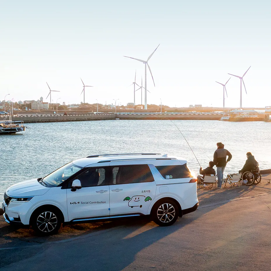 Kia mobility support vehicle parked at a waterfront near people using wheelchairs, with wind turbines in the background