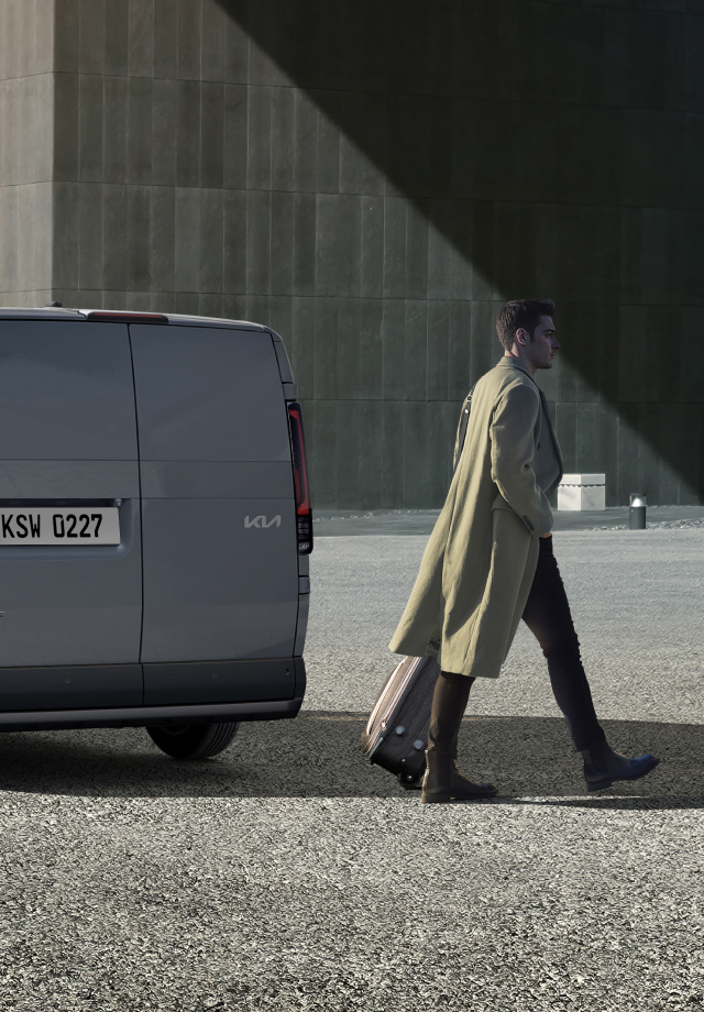 Rear three-quarter view of the Kia PV5 Cargo parked near a building with a man walking by
