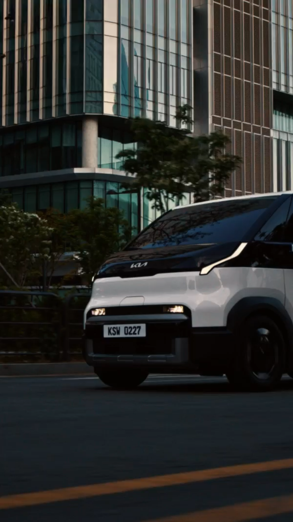 A white Kia PV5 electric passenger van drives through an urban setting as families, children, older adults, friends, and large dogs enter through its wide sliding doors, showcasing the spacious, versatile, and accessible interior of Kia’s electric PBV.