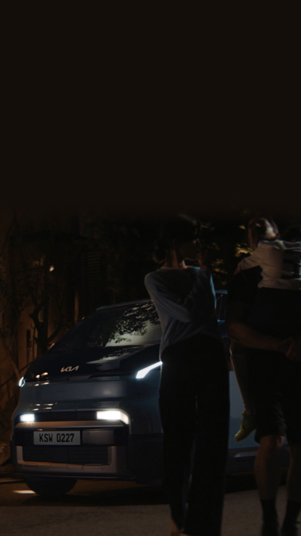 The Kia PV5 Passenger at night with headlights on, as a family stands in front of the vehicle