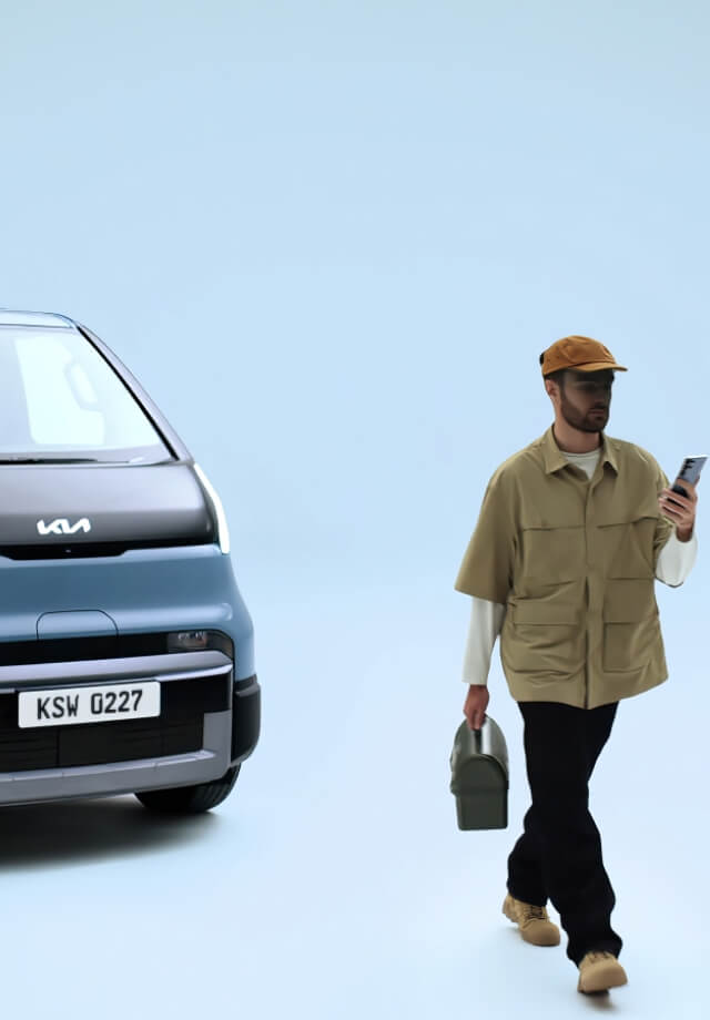 Three-quarter front view of the Kia PV5 Passenger with a man walking while holding a phone and lunchbox