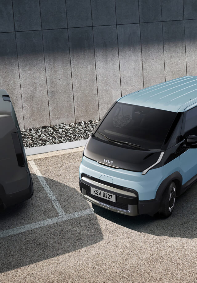 The Kia PV5 Passenger exiting parallel parking space, in a lot with two other vans