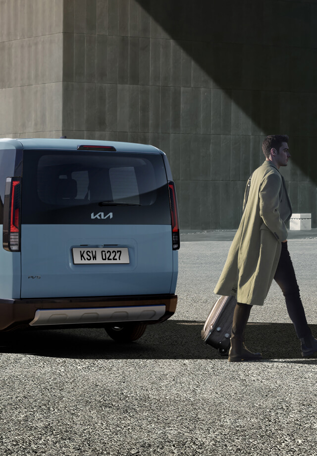 Rear three-quarter view of the Kia PV5 Passenger parked near a building with man walking by