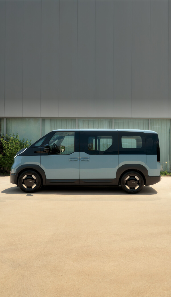Front view of the Kia PV5 Passenger connected to charging station with man sitting outdoors beside camping setup