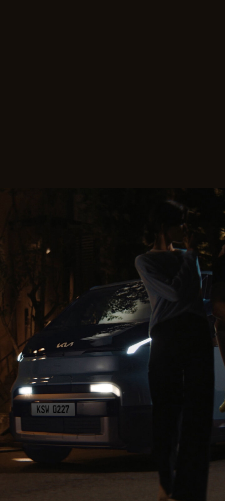 The Kia PV5 Passenger at night with headlights on, as a family stands in front of the vehicle