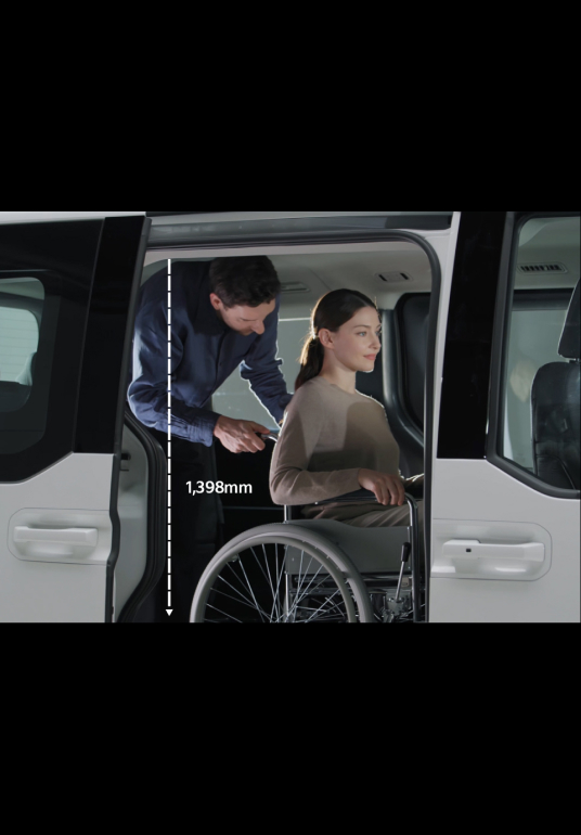 Ein Rollstuhlfahrer steigt mit Unterstützung einer weiteren Person im Fahrzeuginneren durch die Seitentür in den Kia PV5 WAV ein.