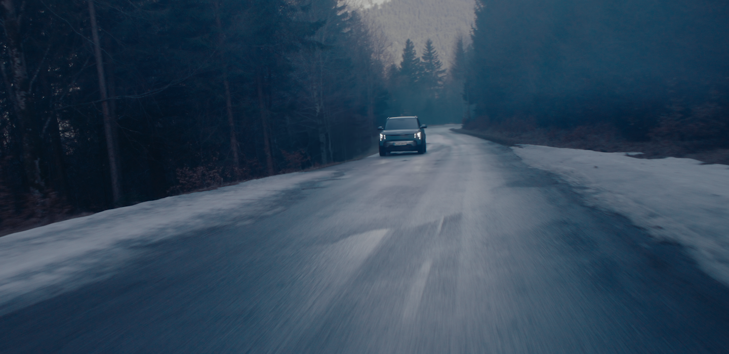 Dark, atmospheric view of a Kia vehicle driving toward the camera on a wet forest road at dusk, headlights on, with patches of snow along the roadside.