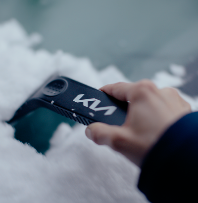 Close-up of a hand using a Kia-branded ice scraper to clear snow and ice from a car windshield.