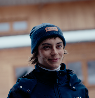 Person smiling outdoors wearing a Kia-branded beanie and navy jacket, with a wooden lodge in the background.