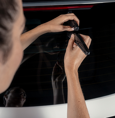 Close-up of a technician's hands installing a small component onto a vehicle's rear window, reflected against the glass.