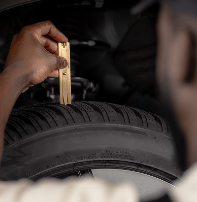 A technician in overalls inspecting a wiper blade component under an open car hood, with a turbocharged engine visible.
