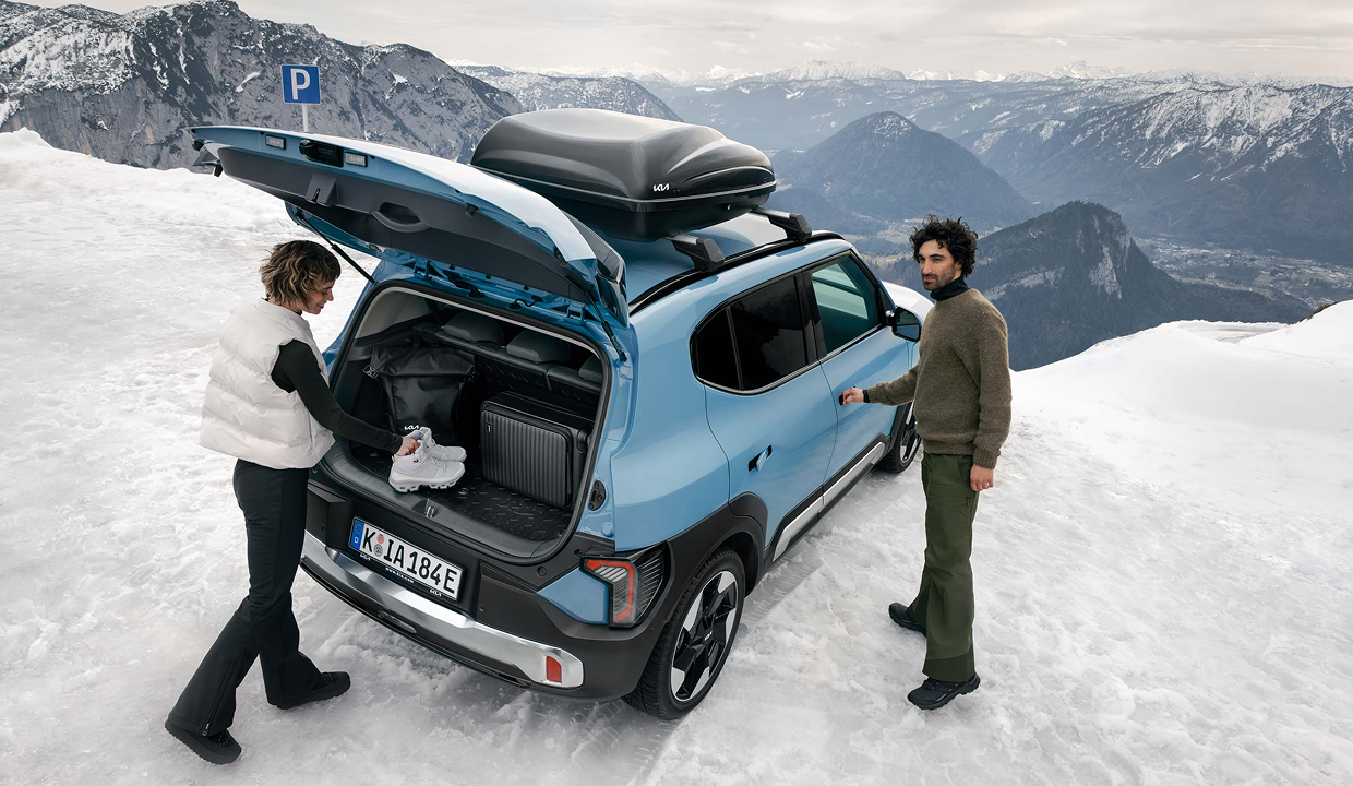 Two people loading luggage and shoes into the open trunk of a blue Kia SUV with a rooftop cargo box, parked on a snowy mountaintop with alpine scenery behind them.
