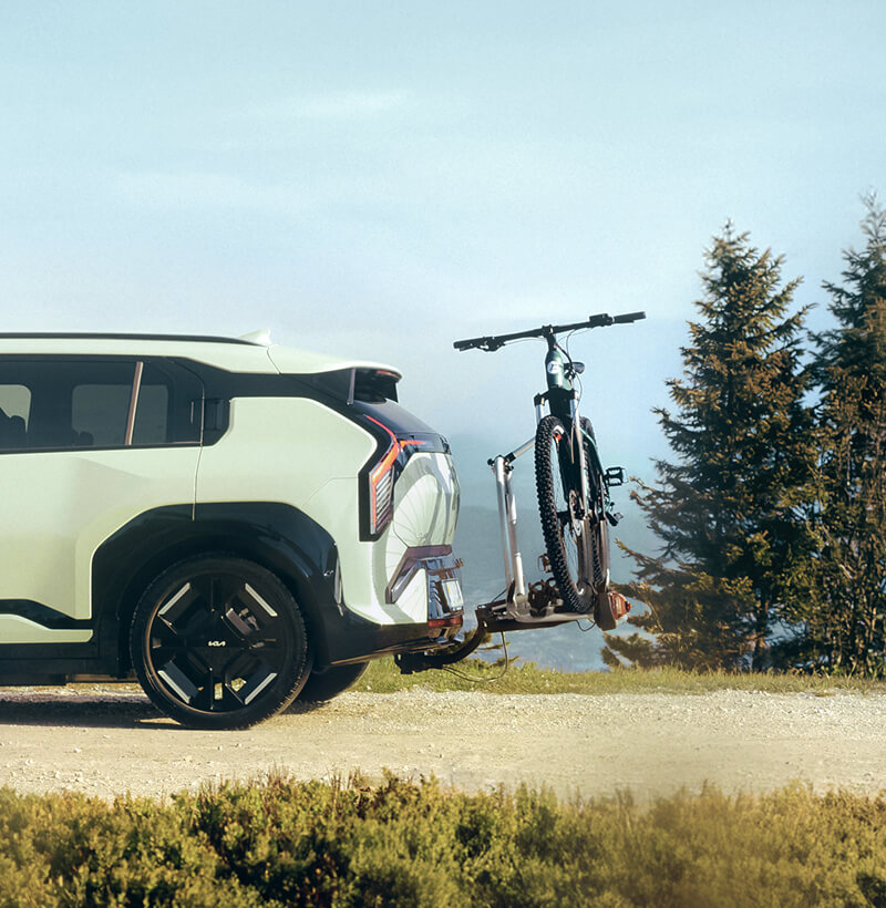 Kia EV3 mit Kia Original-Anhängerkupplung-Fahrradträger, der ein Fahrrad in einer malerischen Außenumgebung transportiert.
