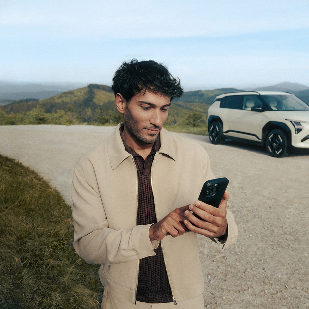 Kia customer stands in front of his Kia EV3 and contacts the Kia Roadside Assistance on his mobile phone.
