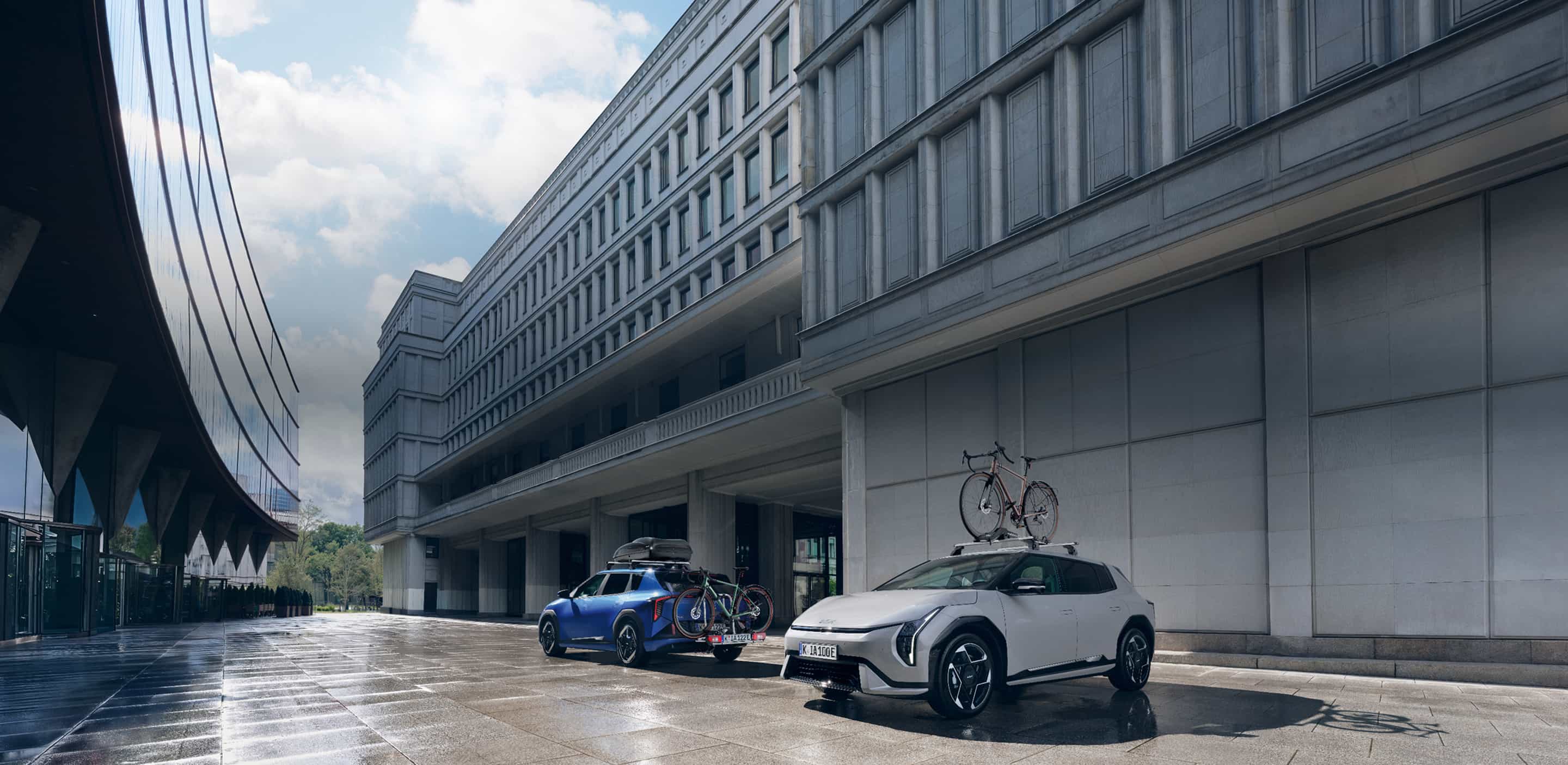 Two Kia EVs equipped with a Kia Roof Box and Bike Carrier Plus park in front of a building.