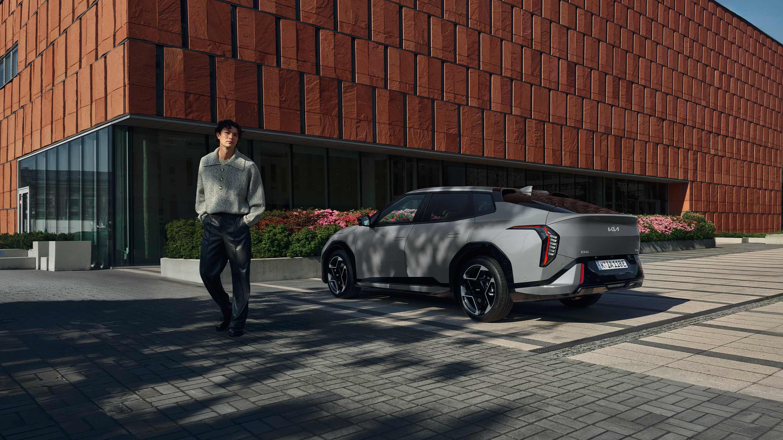 Kia customer confidently leaving his Kia EV4 parked in front of modern city office building on sunny summer day.