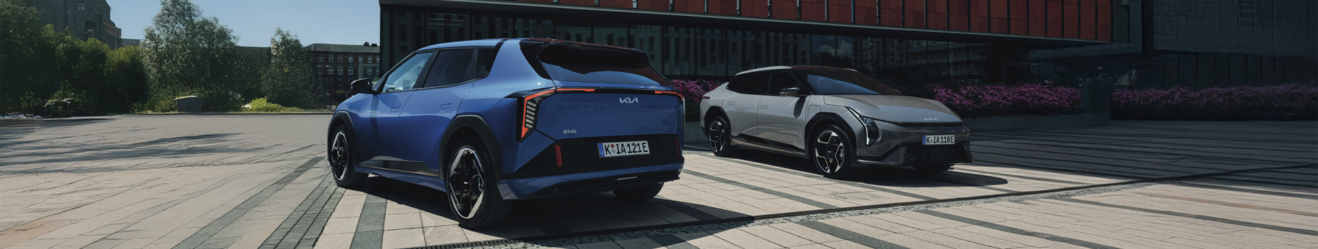 A side profile of the Kia GT-Line S EV6 in Wolf Grey, with a man sat in the passenger seat his legs out of the car. The EV is parked in an area with stones on the floor, and cylindrical cooper panelled building behind.