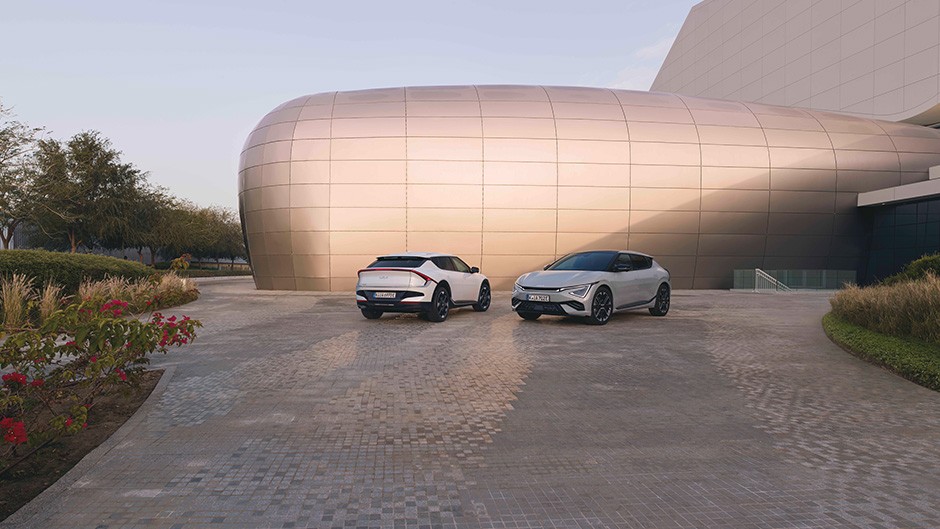 Two Kia GT-Line S EV6’s outside a bricked building. The Wolf Grey EV6 is parked next to the building, whilst the White Pearl EV6 is driving past the parked car.